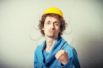 Curly man in a yellow helmet shows the evil revenge, blow fist. On a gray background.