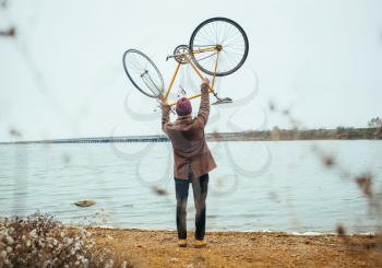 a young man raise over his head bicycle near the lake