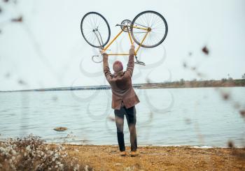 a young man raise over his head bicycle near the lake