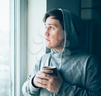 Relaxed young man in hood drinking coffee at home, near the window