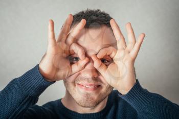 A man looks through the glasses of his fingers. On a gray background.