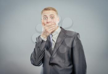 Portrait of afraid businessman wearing suit covering his mouth with his hand over grey background.
