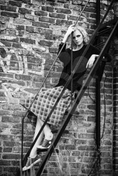 Grunge fashion: cute young girl (informal model) in checkered skirt and jacket sitting on the ladder. Black and white 