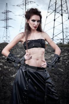 Informal fashion: the beautiful slim young gothic girl dressed in black leather skirt and gloves. Outdoor portrait in field near power line towers