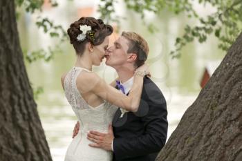 Just married couple kissing between tree trunks and pond
