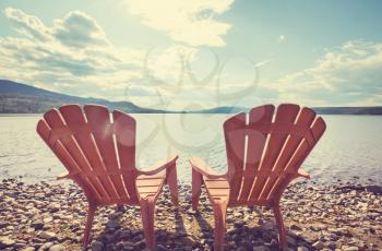 Two wooden  chairs on the shore of a lake facing the water. 