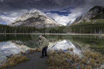 A photographer frames up a reflective image in the stillness of Buller Pond in Alberta Canada's Rockie Mountains.