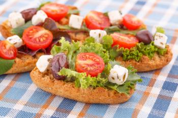 Sandwiches with rusks, vegetables, olives and feta cheese on colorful tablecloth.
