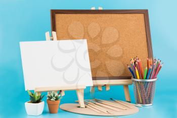 Two wooden easel with blank canvas, cork board, brushes and colorful pencils on blue background.