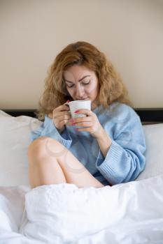 Woman in blue sweater with the cup of tea sitting in the bed.
