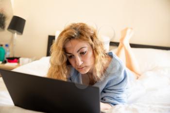 Woman in blue sweater with laptop lying in the bed.