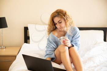 Woman in blue sweater with the cup of tea and laptop sitting in the bed.