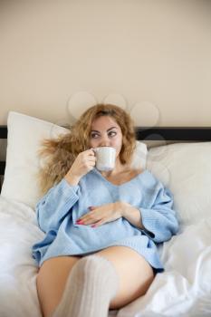 Woman in blue sweater with the cup of tea lying in the bed.