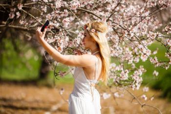 Young beautiful romantic blonde woman in white dress taking a selfie in blooming garden.