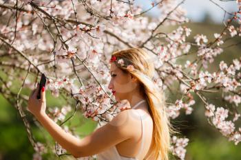 Young beautiful romantic blonde woman in white dress taking a selfie in blooming garden.