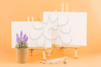 Two wooden easels with blank canvases, brushes and lavender flowers on yellow background.