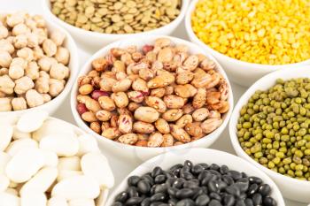 The collection of different beans and peas in the ceramic bowls on a white background.
