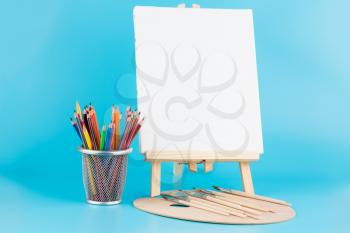 Wooden easel with blank canvas, brushes and colorful pencils on blue background.