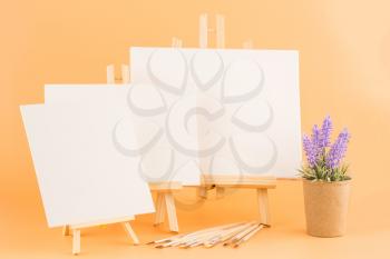 Three wooden easels with blank canvases, brushes and lavender flowers on yellow background.
