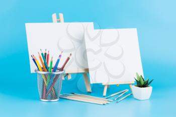 Two wooden easels with blank canvases, brushes, pencils and plant on blue background.