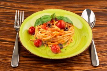Pasta spaghetti Napoli or Napolitana on green plate with fork and spoon on brown background. Italian cuisine.