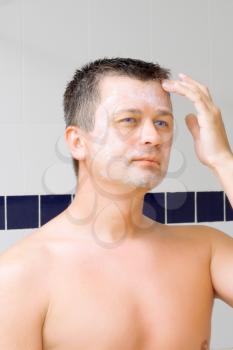 A young man puts on face cream mask in bathroom.