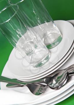 Pile of white plates, glasses with forks and spoons on silk napkin. Green background