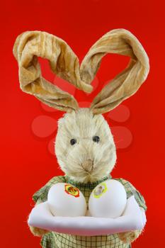 Straw rabbit with eggs in hands on red background.