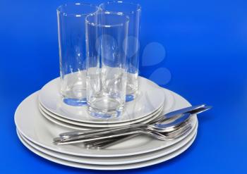Pile of white plates, glasses with forks and spoons on silk napkin. Blue background