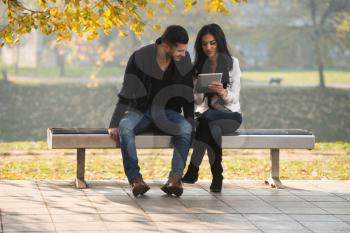 Beautiful Young Couple Sitting On Bench In The Park On A Beautiful Autumn Day - They Are Using Internet Via Digital Tablet