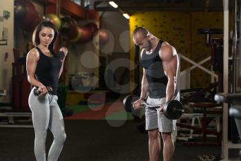 Strong Young Couple Doing Exercise For Biceps With Dumbbells And Barbell In The Gym