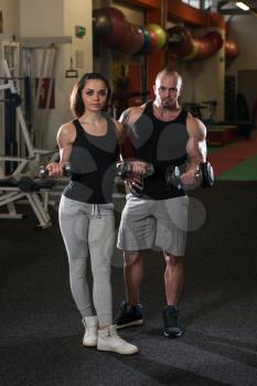 Strong Young Couple Working Out With Dumbbells For Biceps In The Gym With Exercise Equipment
