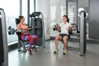 Strong Young Couple Doing Exercise For Biceps On Machine In The Gym