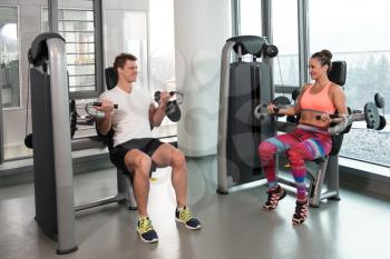 Strong Young Couple Doing Exercise For Biceps On Machine In The Gym