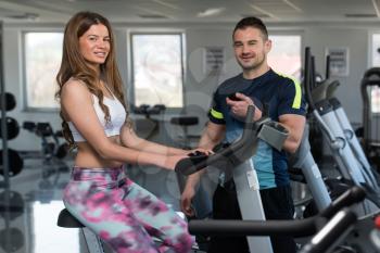 Young Woman In The Gym with Personal Trainer - Exercising Her Legs Doing Cardio Training On Bicycle