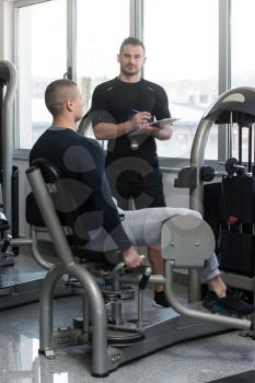 Personal Trainer Takes Notes While Young Man Exercise Legs On Machine In The Gym