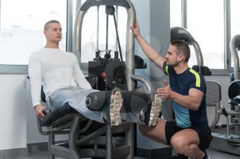 Personal Trainer Showing Young Man How To Train Legs On Machine In The Gym