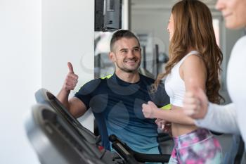 Personal Trainer Showing Ok Sign To Client - Group Of People Exercising On Elliptical Walker In Gym Or Fitness Club