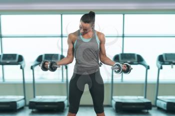Young Woman Exercising Biceps With Dumbbells In The Gym And Flexing Muscles - Muscular Athletic Bodybuilder Fitness Model Doing Dumbbell Concentration Curls