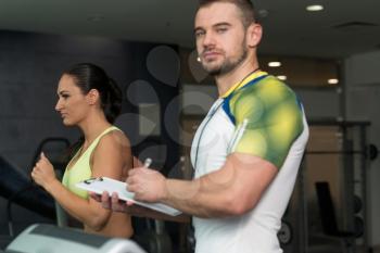 Woman With Fitness Trainer Running On Treadmill In Gym Or Fitness Club While Personal Trainer With Clipboard Watching Her