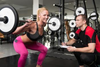 Young Woman Exercise Legs Squat  In The Gym While Personal Trainer Helps Out