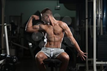 Handsome Geek Man Sitting Strong In The Gym And Flexing Muscles - Muscular Athletic Bodybuilder Fitness Model Posing After Exercises