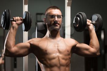 Handsome Man Wearing Eyeglasses Working Out Shoulders With Dumbbells In A Dark Gym