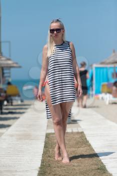 Young Blonde Woman In Dress And Sunglasses Walking On Beach
