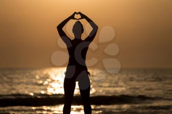 Beautiful Female Model Enjoying Sunset and Making Heart Sign on Sun at Seaside - Calm Water Reflects Silhouette of Woman - Sun Goes Under Horizon