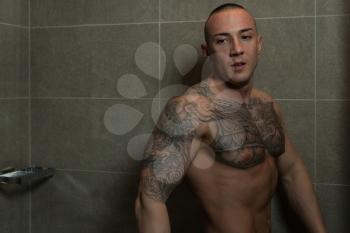 Young Good Looking and Attractive Tattoo Man With Muscular Body Wet Taking Shower in Bath With Black Tiles in Background