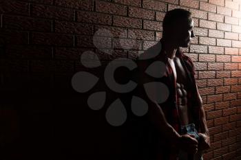 Healthy Young Man Standing Strong In The Gym And Flexing Muscles While Wearing Plaid Shirt - Muscular Athletic Bodybuilder Fitness Model Posing After Exercises On Wall of Bricks