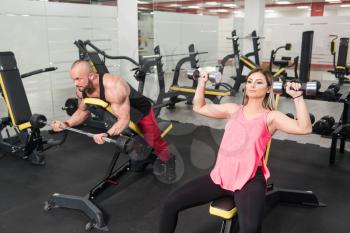 Strong Young Couple Working Out With Dumbbells And Barbell For Biceps And Shoulders In The Gym With Exercise Equipment