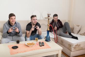 Three Young Gamers Sitting Together On Sofa And Playing Video Games At Home