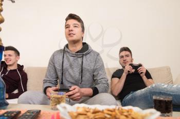 Three Young Gamers Sitting Together On Sofa And Playing Video Games At Home